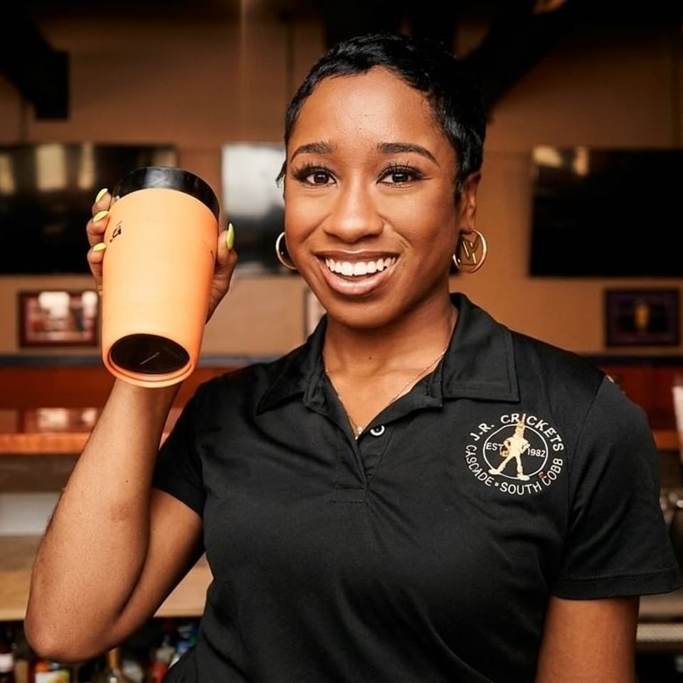 Smiling woman in a black J.R. Crickets polo shirt holding up an orange travel mug.