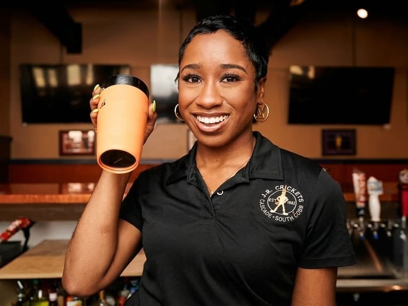 Smiling woman in a black J.R. Crickets polo shirt holding up an orange travel mug.
