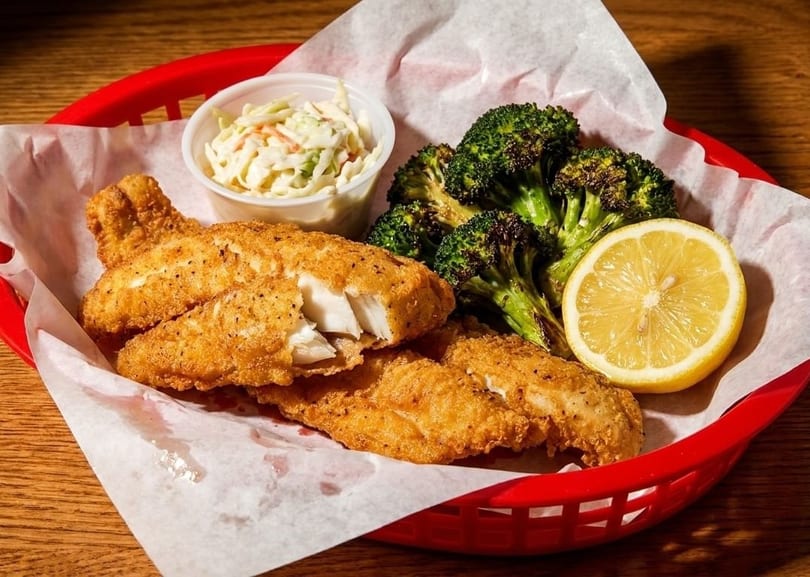 Fried fish fillets with coleslaw, roasted broccoli, and a lemon wedge in a red basket.